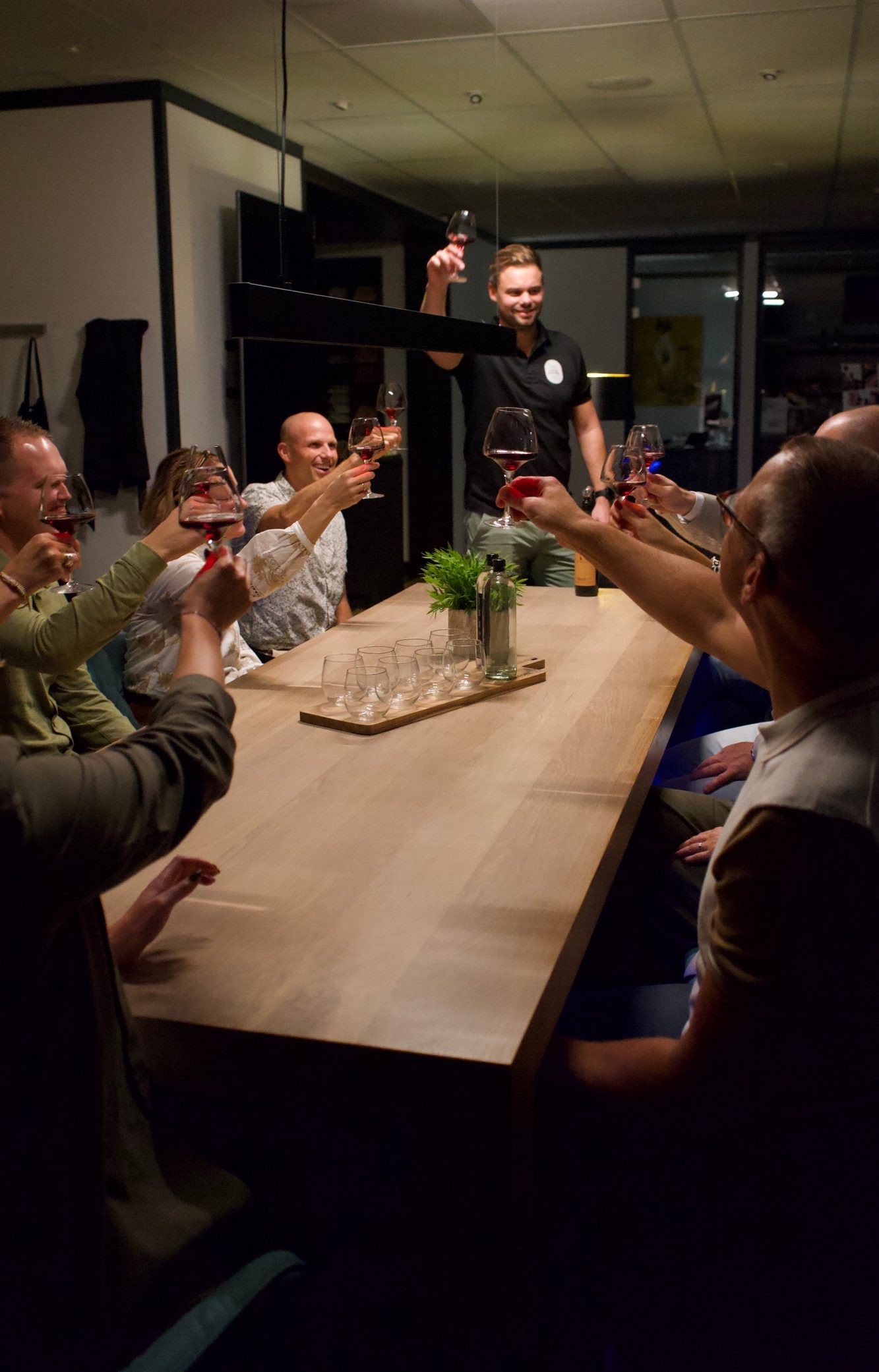 Gezellige wijnproeverij aan huis waar gasten proosten met een glas wijn aan een lange tafel.
